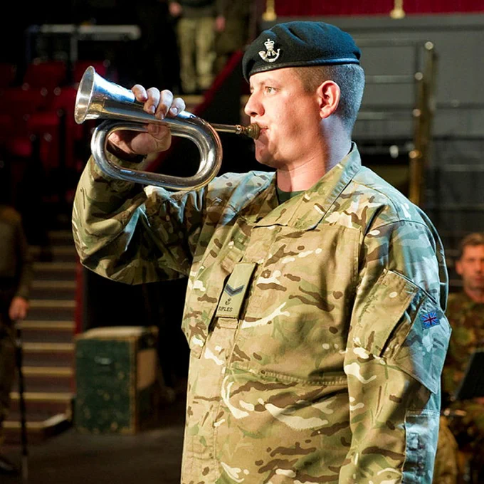 A person in camouflage military uniform and a black beret plays a silver bugle indoors, standing in profile with rows of red theatre seats blurred in the background.