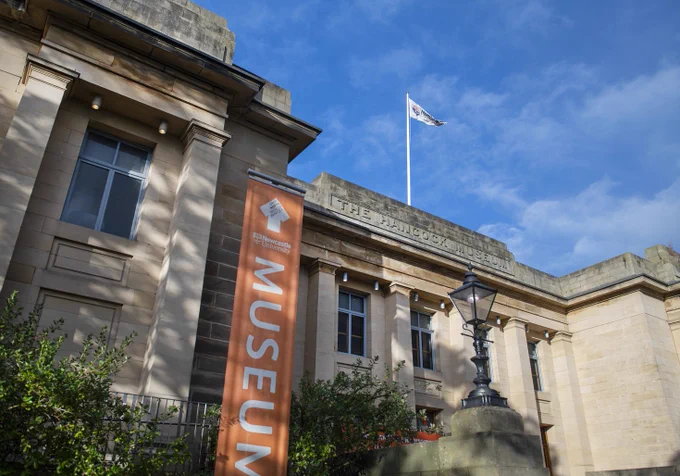 The exterior of a large stone building, with 'The Hancock Museum' carved into the stone above the entrance. There is a large vertical sign which reads 'Museum' in white text on an orange background.