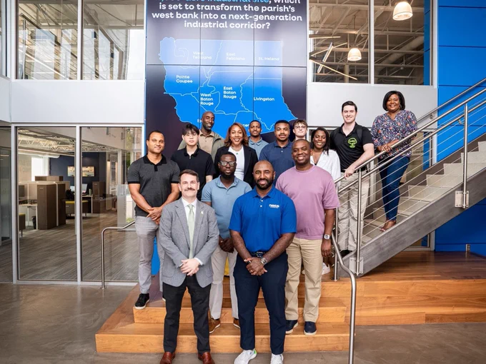 A group of thirteen people pose for a photo on and in front of a staircase inside a modern office with a map and informational sign in the background.