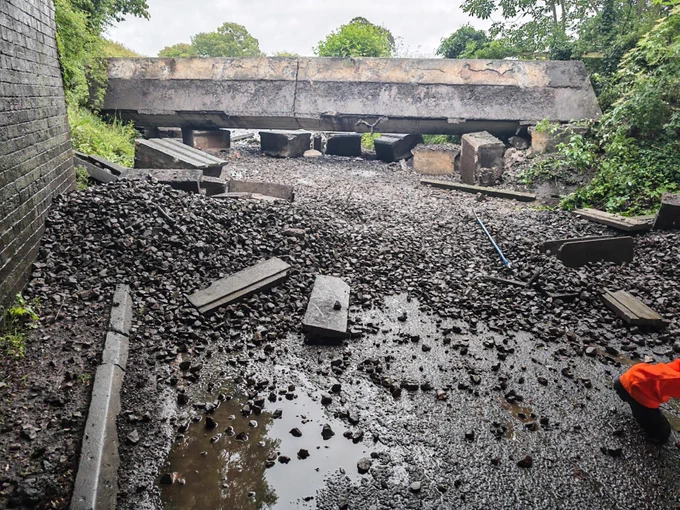 A collapsed rail bridge is seen with debris and rocks scattered across a muddy ground. 