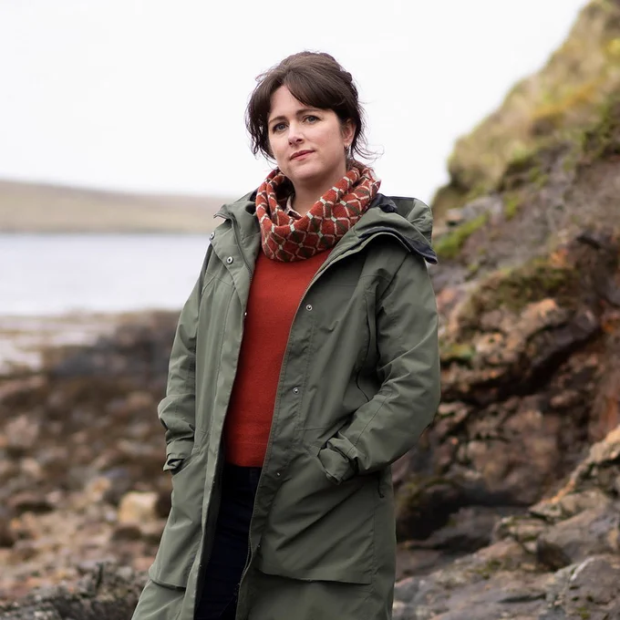 Alison O’Donnell in character as Tosh. Wearing a long coat, a jumper and scarf, she stands on a rocky beach beside water. 