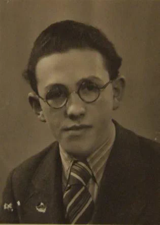 Vintage black-and-white portrait of a young man wearing round glasses, a suit, and a striped tie, with a serious expression.
