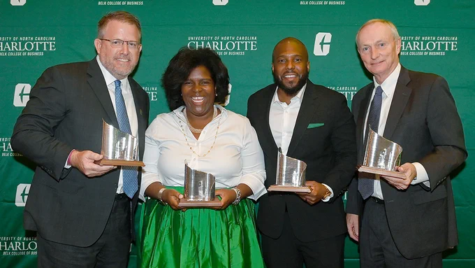 Four smiling individuals stand in front of a green UNC Charlotte Belk College of Business backdrop, each holding an alumni award.
