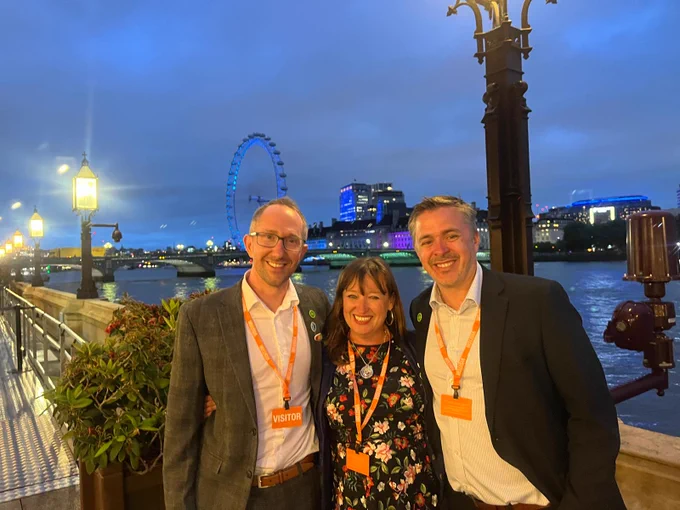 Three people wearing visitor badges pose on a riverside walkway at dusk with the illuminated London Eye in the background.