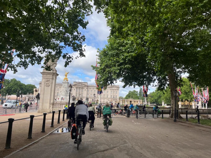 Cyclists riding along a tree-lined path near Buckingham Palace with Union Jack flags fluttering in the breeze.