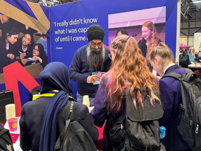 A group of individuals is gathered around a table at a booth displaying an educational event. One person is interacting with the others, demonstrating or explaining something. On the backdrop is a quote from a student: "I really didn’t know what I was capable of until I came." The scene takes place in a large indoor arena with other booths visible in the background.