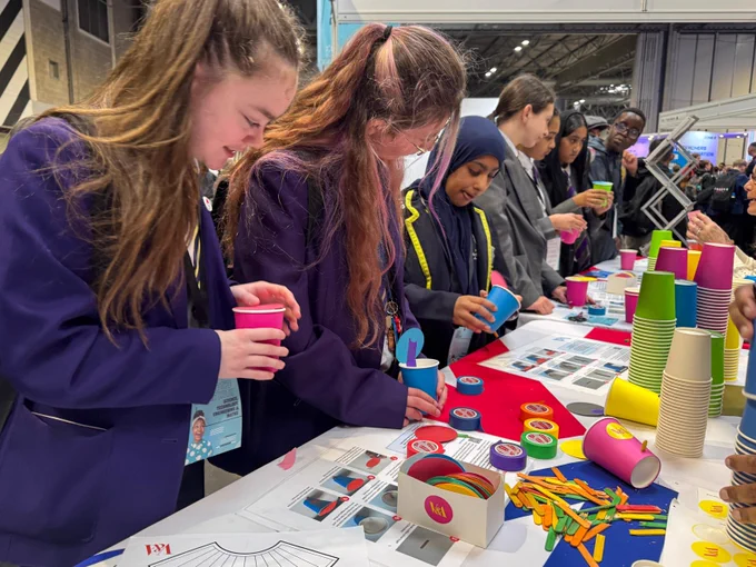 A group of people in school uniforms are engaging in a hands-on activity at a table filled with colorful paper cups, stickers, and crafting materials. The setting appears to be a lively event or workshop.