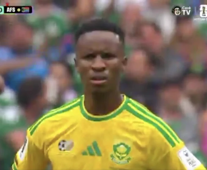 A player in a yellow soccer uniform looks concerned during a match, with a blurred crowd in the background suggesting a tense atmosphere.