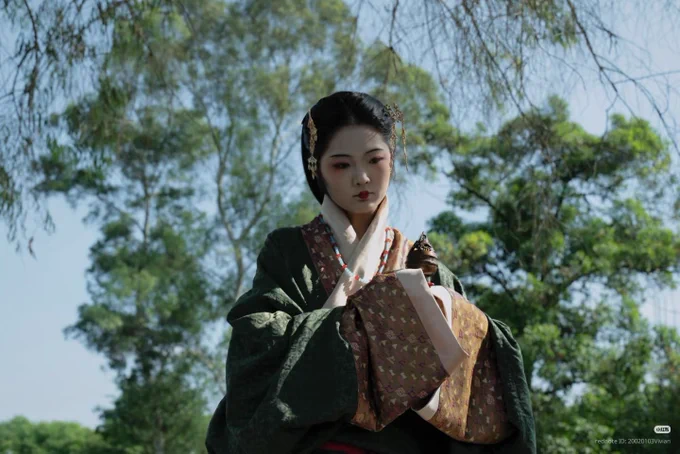 Woman in Han dynasty hanfu, dark green with copper and cream borders. Hair pinned back. In a park.