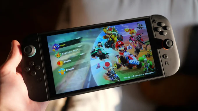 A hand holds a dark gray Nintendo Switch console displaying the character select screen of Mario Kart World, with options for Race, Knockout Tour, Battle, and Friends.