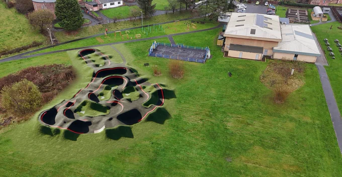 An aerial view of a park featuring a new pump track with red-lined edges and dark dips, surrounded by grass.