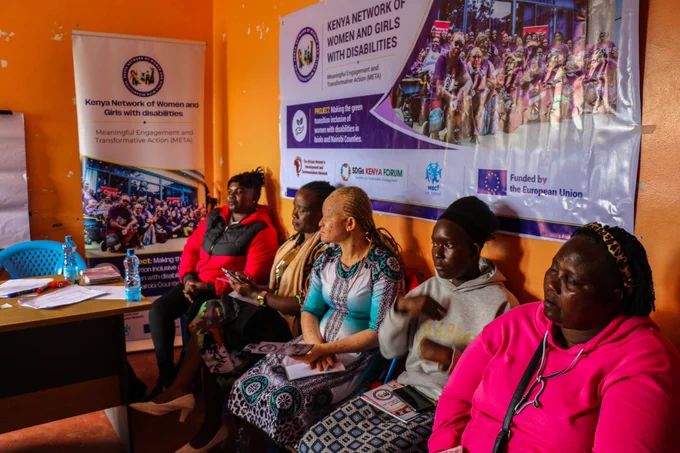 This captures a group of women participating in an event focused on disability inclusion and empowerment.The central focus of the image is a group of women seated. From left to right, the first woman is wearing a red and black jacket.Next to her is a woman with braided hair, wearing a patterned top and a light brown scarf and she is looking to the side.The third woman, also with braided hair, is wearing a blue and black patterned dress and has a notebook and pen on her lap. The woman next to her, wearing a grey hoodie, is looking away from the camera. To the far right, a woman in a bright pink hoodie is visible, with her head tilted slightly. Behind the women, a large banner is displayed. It features text, "KENYA NETWORK OF WOMEN AND GIRLS WITH DISABILITIES," "Meaningful Engagement and Transformative Action (META)," and logos for SDGs Kenya, WECF International, and the European Union.