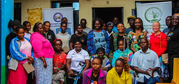 A diverse group of people, many are women with disabilities, gathered together outdoors in front of buildings and banners after the event.They  are standing and sitting in front of a building with yellow and blue painted walls. Several banners are visible, one with text in English that includes "KENYA NETWORK OF WOMEN AND GIRLS WITH DISABILITIES" and another with a logo and text that reads "DISABLED INCLUSION NODE ASSOCIATION". The people are dressed in a variety of colorful and casual clothing, including t-shirts, sweaters, jackets, and skirts. 