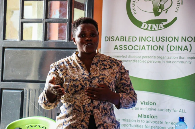 A woman speaks passionately at a podium in front of a banner for the Disabled Inclusion Node Association. The image features a woman with dark skin and short, curly dark hair, speaking with an animated expression. She is wearing a patterned shirt with paisley designs in shades of gold, brown, and dark blue, featuring a V-neckline and buttons down the front. Her left hand is extended slightly outwards, palm open,emphasizing a point, while her right hand rests on her chest, near her heart, with a visible ring on her finger.
