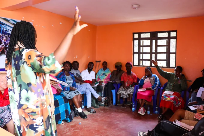This is a discussion taking place in a modest indoor setting. A facilitator stands at the front-left of the room, facing the participants. The facilitator has one hand raised, leading a discussion. Several participants are seated in a circle on plastic chairs, listening attentively.
One participant has raised their hand, suggesting an interactive session.