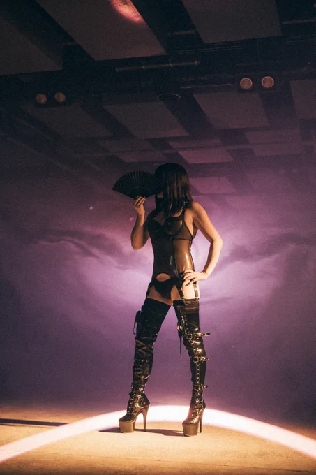 Trans Princess Jezebel is standing in front of a dark purple mist and a round beam of bright light on the ground dressed in a sheer latex corset, black stockings and black thigh-high, strapped, high heeled boots, her long hair flowing down her torso. She has one hand on her left hip and the other holding a fan over her face.