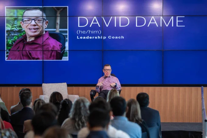Speaker in a wheelchair presents to an audience; large screen behind shows name and title, “David Dame, Leadership Coach.”