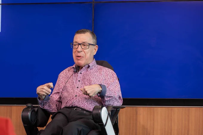 Speaker seated in a wheelchair speaks on stage, gesturing while sharing leadership insights.