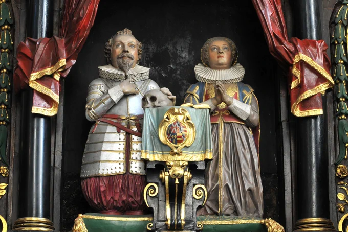 A detailed sculpture depicting Lady Elizabeth Fane and her husband Sir George in historical attire. Sir George wears armor with a ruffled collar and holds a skull, while Lady Elizabeth has a ruffled collar and clasps her hands in prayer. They are both positioned behind a lectern adorned with a colorful coat of arms. Red drapery with gold trim frames the figures.
