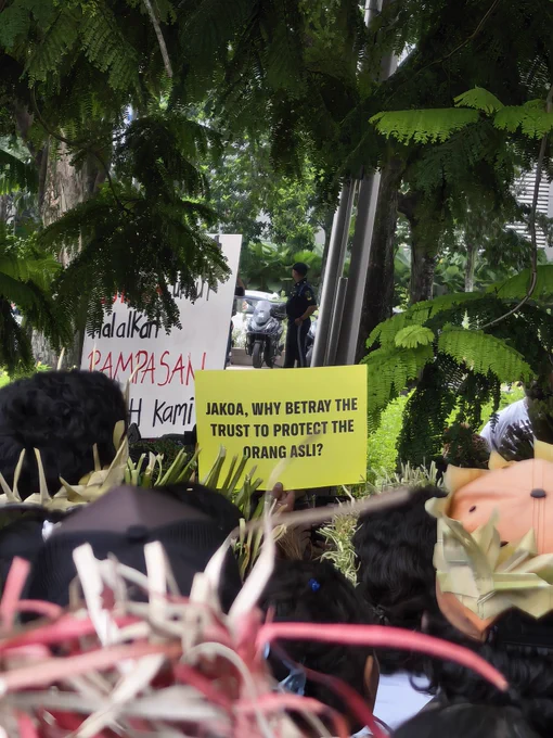 A view from behind a crowd of protesters looking toward a bright yellow sign held aloft under large green trees. The yellow sign reads in bold black capital letters: "JAKOA, WHY BETRAY THE TRUST TO PROTECT THE ORANG ASLI?" In the background, partially obscured by leaves, a larger white sign has red and black handwriting, and a security guard or police officer in a dark uniform stands near a motorcycle parked on a street. Protesters in the foreground wear traditional woven headbands and hats.