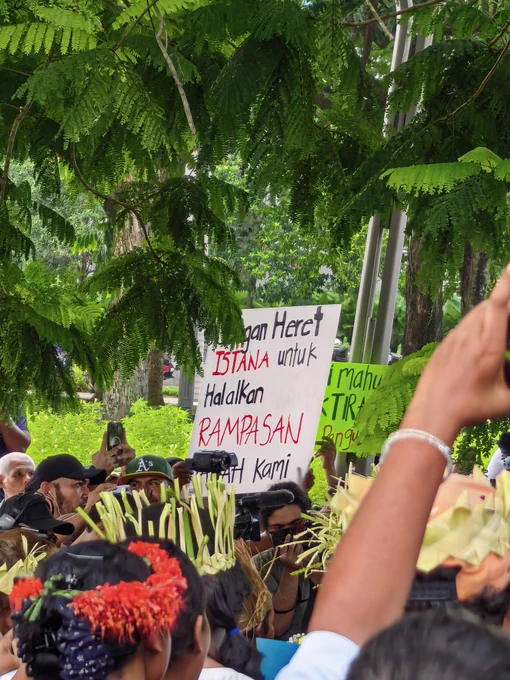 A candid shot taken within a crowded protest under a canopy of green trees. In the center, a large white cardboard sign is held up with bold, handwritten text in red and black ink that reads: "...gan Heret ISTANA untuk Halalkan RAMPASAN [Ta]nah Kami". Other green protest signs are partially visible nearby. People in the crowd are holding up smartphones and a video camera to record the event, and some individuals wear traditional floral and woven leaf headpieces.