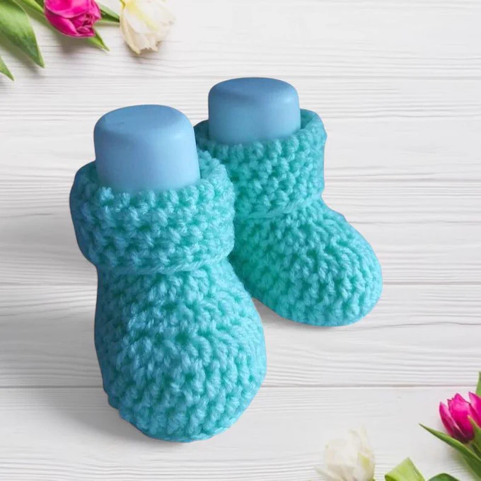 A pair of spring green crocheted baby booties with a textured stitch sit side by side on pale whitewashed wood. Each has a white foot form insert inside to make the booties stand. Fresh tulips in pink, white and yellow with green leaves frame the top left and bottom right corners, creating a bright, delicate and fresh scene.