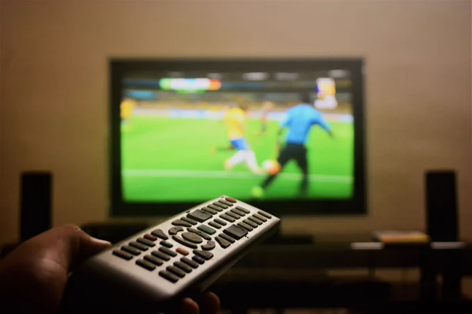 A hand holds a TV remote in the foreground, pointing towards a flatscreen television in a dimly lit room. On screen, two footballers compete for the ball on a green pitch.