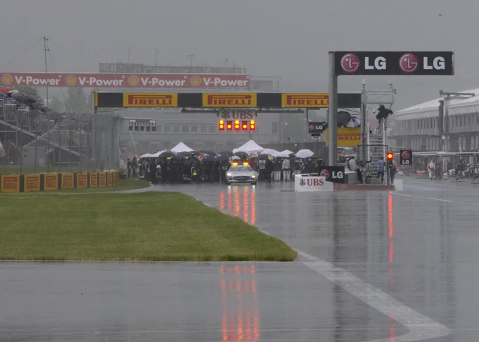 Shot from a distance at Virage Senna in heavy grey gloom, the red and yellow lights of the gantry and pit exit streak downwards as teams and drivers are gathered on the pit straight under umbrellas and gazebos. The safety car sits at the front, white and yellow lights glowing. Rainer Schlegelmilch