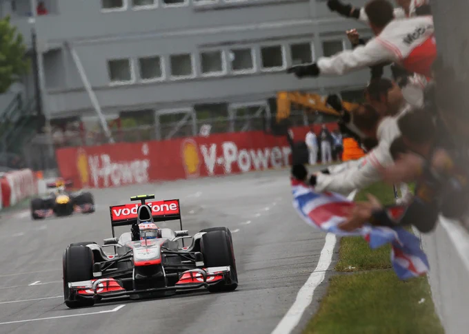 Shot from pit wall looking back down the straight, Jenson Button waves his hand out of the cockpit of his chrome and red McLaren MP4/26 as members of the McLaren team lean through the fence to celebrate the win. Daniel Kalisz.