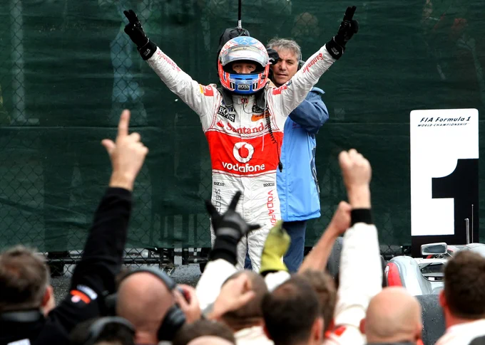 In the centre of frame, Jenson Button stands with his arms raised and eyes wide, wearing a white and red Vodafone branded race suit and blue, white and red crash helmet with the visor open. He is filmed from behind by a camera man, while in the bottom of frame, wave their arms in the air in celebration. Andy Hone.