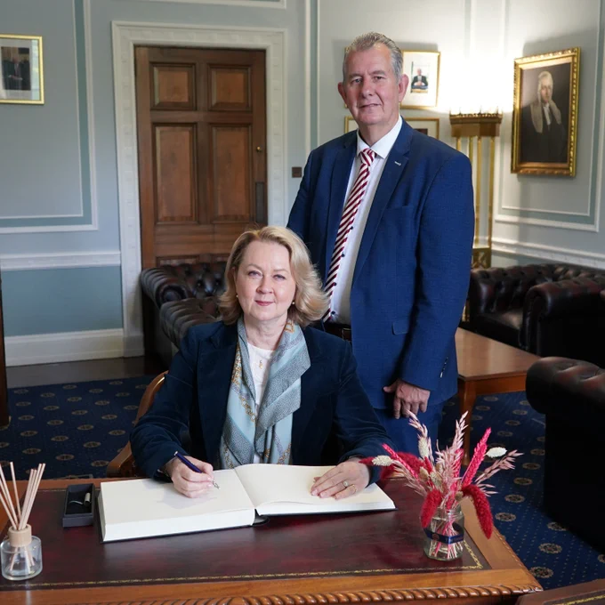 The Speaker and Ms Leena Linnus in formal business attire are pictured in an elegant office setting with panelled walls, framed portraits, and leather furniture. Ms Leena Linnus is seated at a wooden desk in the foreground, signing a large open book with a pen. The desk holds a pen stand, a small container with sticks, and a vase with red and cream dried flowers. A second person stands just behind and to the right of the seated person, facing the camera. Soft natural light illuminates the scene, and a wooden double door is visible in the background.