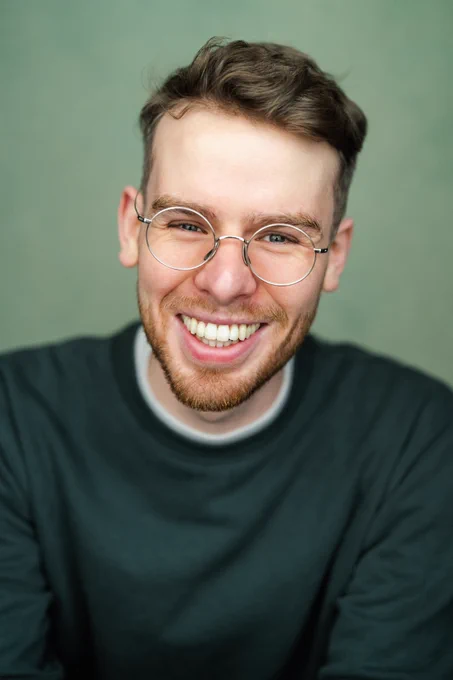 A photo of author Ollie George Clark. He is white, young, bearded, wears glasses and is smiling into the camera. 