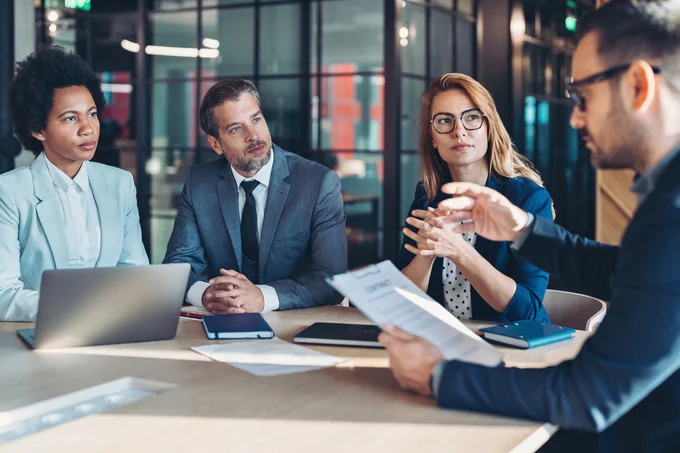 Four professionals engaged in a business meeting around a table with laptops and documents in a modern office setting.