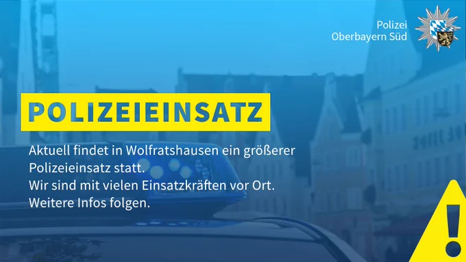 Die Grafik mit blauem Overlay zeigt das Blaulicht eines Streifenwagens. Im Hintergrund ist eine Häuserreihe zu erkennen.  In der Mitte links steht in einem gelben Textbalken "Polizeieinsatz" und in weißer Schrift darunter "Aktuell findet in Wolfratshausen ein größerer Polizeieinsatz statt. Wir sind mit vielen Einsatzkräften vor Ort. Weitere Infos folgen". Oben rechts ist die Wort-Bild-Marke der Polizei Oberbayern Süd abgebildet. Unten rechts steht in weißer Schrift "Mehr Informationen im Beitrag". Unten rechts im Eck befindet sich ein gelbes Warndreieck.