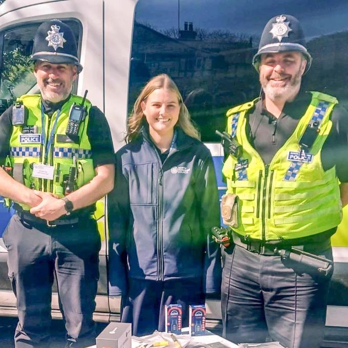 Two police officers and a council employee smile for a photo in front of a van, with items on a table.