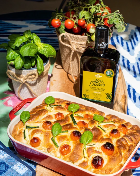 Focaccia con tomates cherry y hojas de albahaca en fuente roja; albahaca en maceta y botella de aceite de oliva.