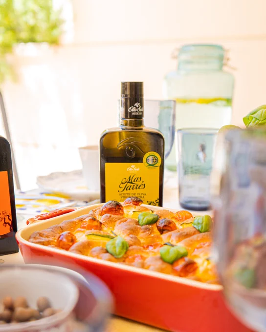 Plato horneado con tomates cherry y hojas de albahaca, frente a una botella de aceite de oliva y vasos.
