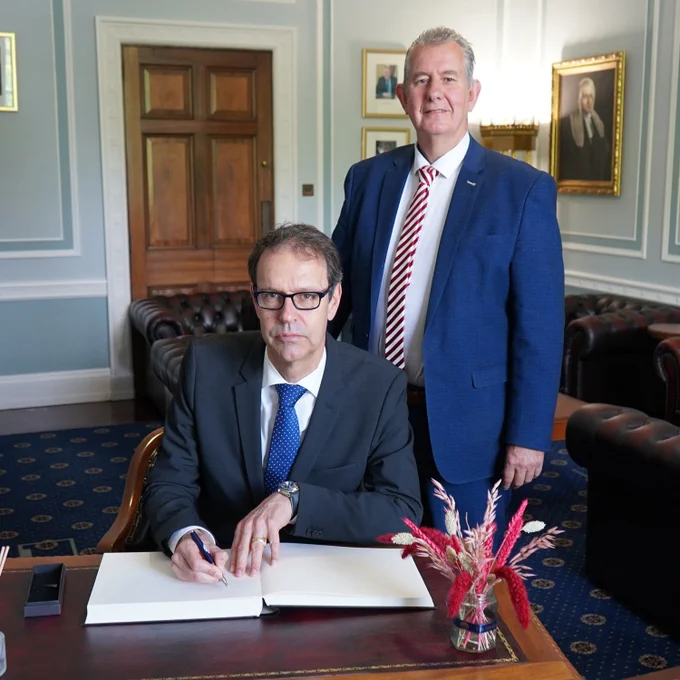 Mr Dominique Paravicini, Ambassador of Switzerland to the UK, sits at a desk signing an open guestbook with a pen, while Edwin Poots MLA, Speaker of the Northern Ireland Assembly, stands beside him. Both are dressed in business attire in a formal office with wood-paneled doors, framed portraits, leather seating, and a small arrangement of red and beige flowers on the desk in the foreground.