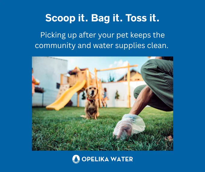 A person crouches on grass, using a plastic bag to pick up after a dog. The dog is sitting in front with a playground slide visible in the background. The image includes the text: "Scoop it. Bag it. Toss it. Picking up after your pet keeps the community and water supplies clean." Opelika Water logo is at the bottom.