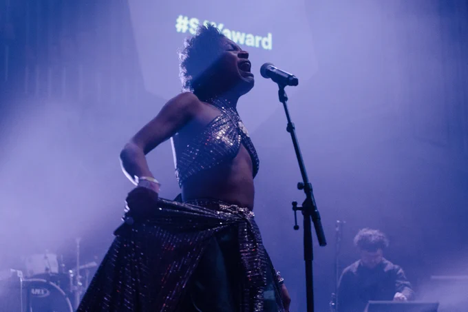 Beldina stands on-stage and sings into a microphone. The lighting is purple. 