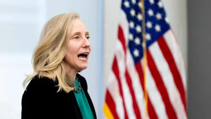 Governor Spanberger, a midforties woman with blond hair, wearing a black suit jacket, speaking in front of an American flag