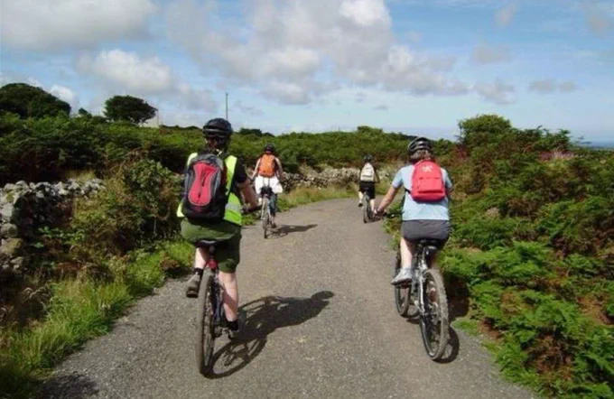 Four cyclists riding on a gravel path through green countryside under a cloudy blue sky.