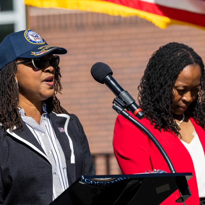 The photo shows two individuals at a podium outdoors. One person is speaking into a microphone, wearing a dark jacket, striped shirt, and a cap. The other person is standing beside them, wearing a red jacket. An American flag is partially visible in the background.