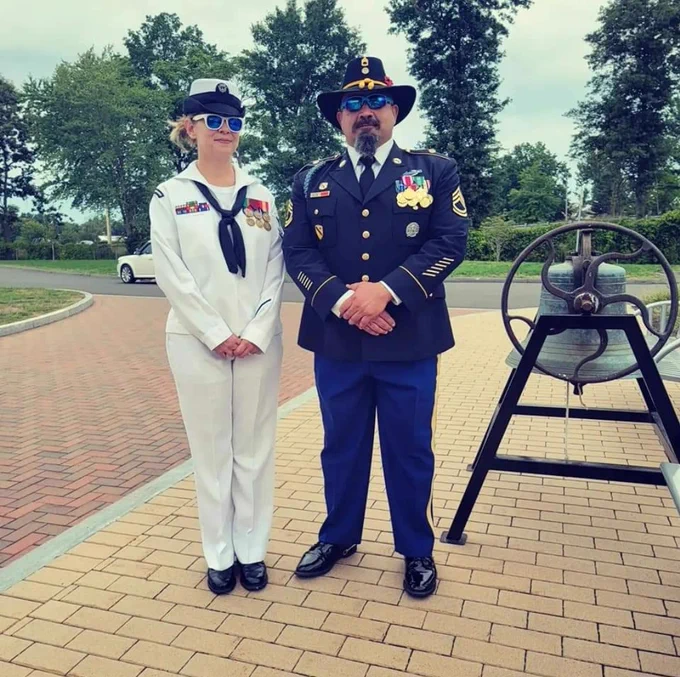 A person in a white naval uniform stands beside someone in a blue military uniform with a black hat. They are in front of a bell with greenery and trees in the background.