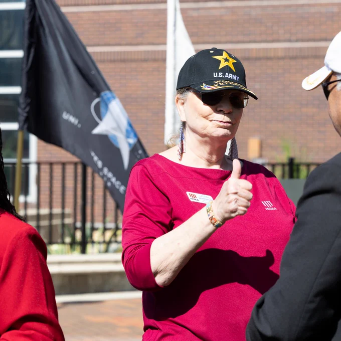 A woman wearing a U.S. Army cap gives a thumbs up to someone facing her. A flag waves in the background.