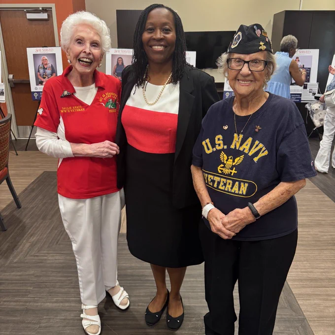 Three smiling individuals are standing together indoors. The person on the left wears a red shirt with a badge, the one in the middle is in a black and red outfit, and the person on the right wears a U.S. Navy Veteran shirt and a cap. There are posters and people visible in the background.