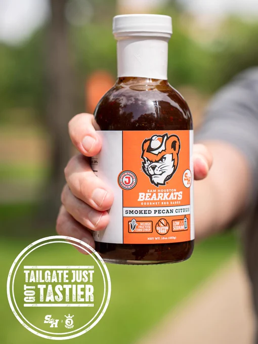 A person holding a bottle of "Sam Houston Bearkats Smoked Pecan Citrus" barbecue sauce. The label features the Bearkat mascot. 