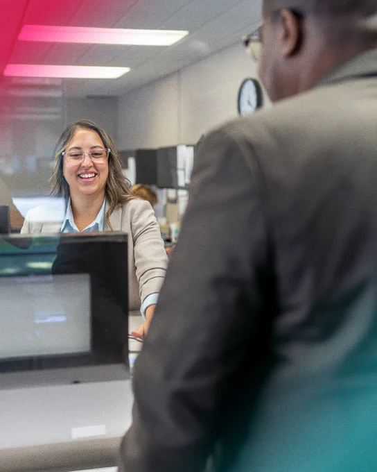 Alt text: A smiling student wearing glasses and business attire speaks with another person across a reception desk in an office setting, creating a welcoming and professional atmosphere.