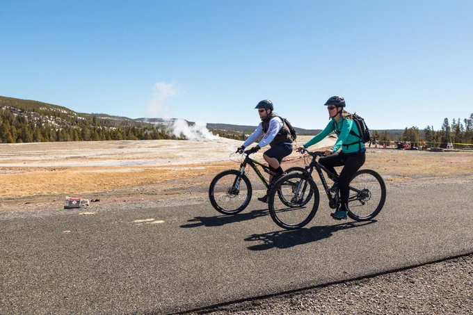 Two people riding bicycles on a paved surface near a thermal feature on a summer day
