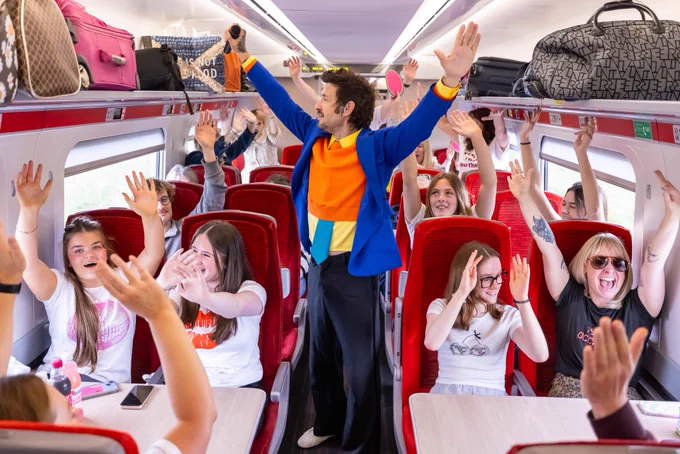 Sean Mac performing his Harry Styles show on an LNER Azuma. Passengers are sitting holding their arms raised.
